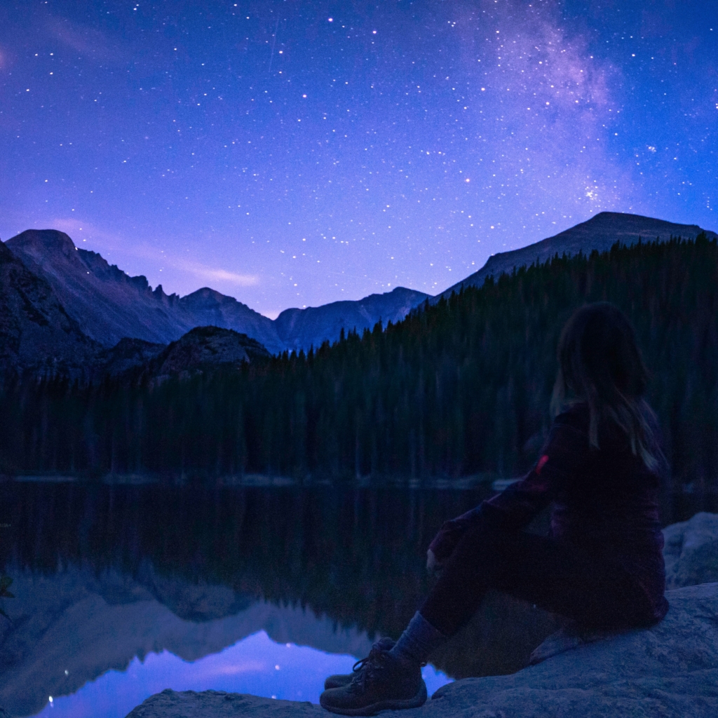 Scorpio - on a hike - woman sitting on rock at lake in wilderness at night and looking at stars