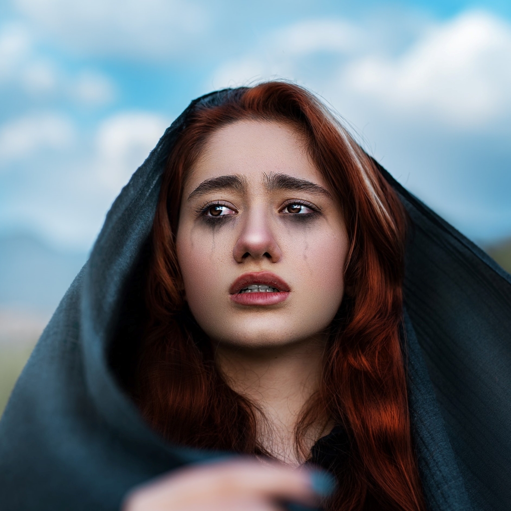 Gemini - professional mourner - redhead woman with large black hood with makeup appearing as though she is crying