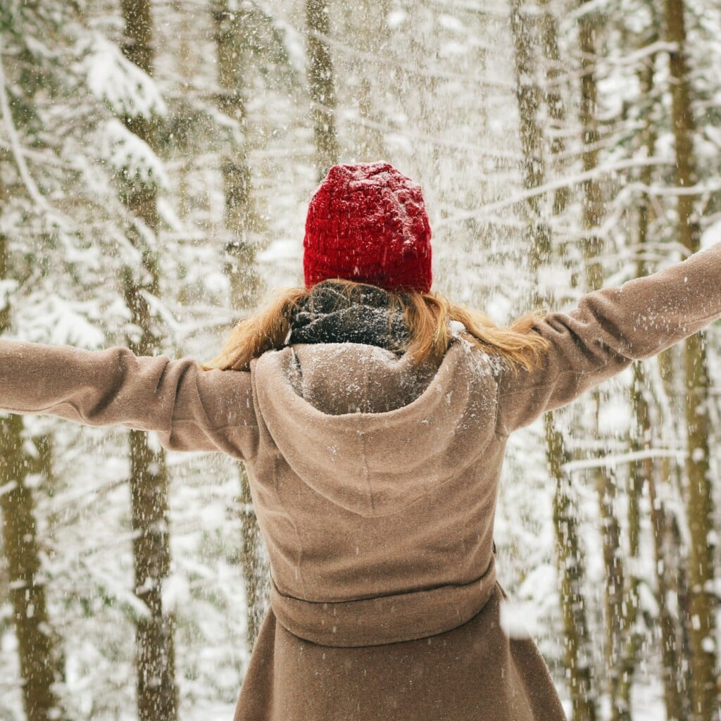 6 ways to relish the cold weather, fire signs! - woman standing in snowy forest with arms outstretched and wearing a camel coat and red winter hat
