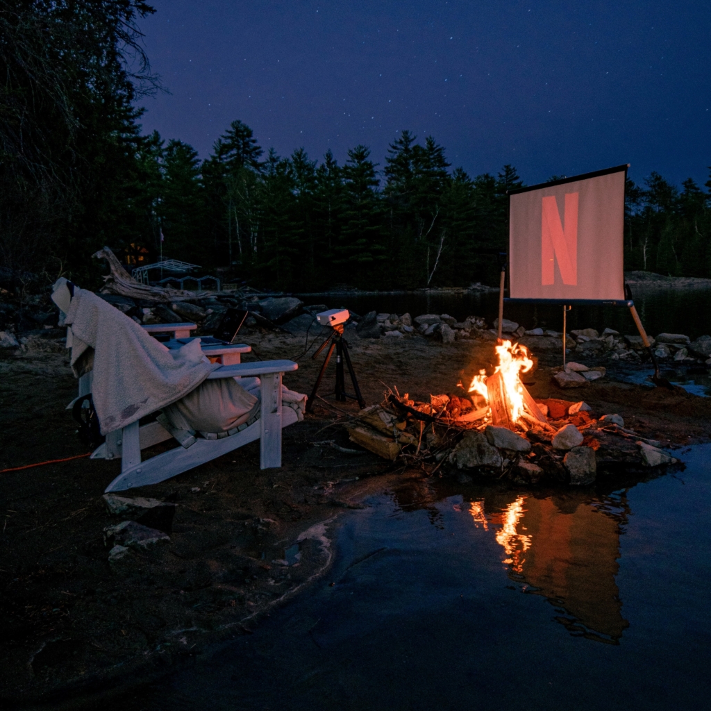 Throw a comfort movie marathon - outdoor chairs with blankets facing a projector with Netflix loading logo and nearby firepit
