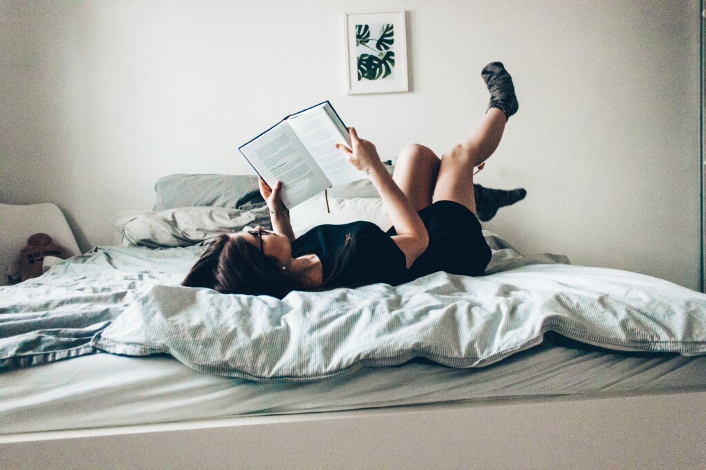 Capricorn - dystopian- woman lying on back in bed with feet up in the air and reading a book
