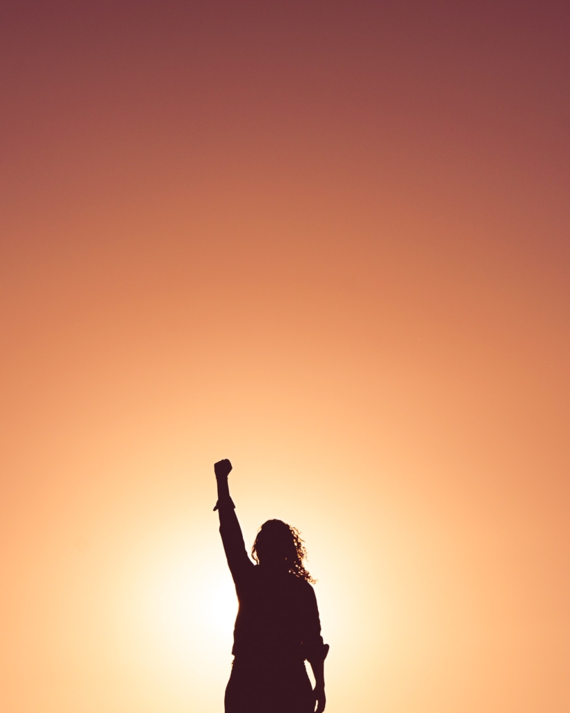 Silhouette of woman standing with one fist in the air against sunset colored background 