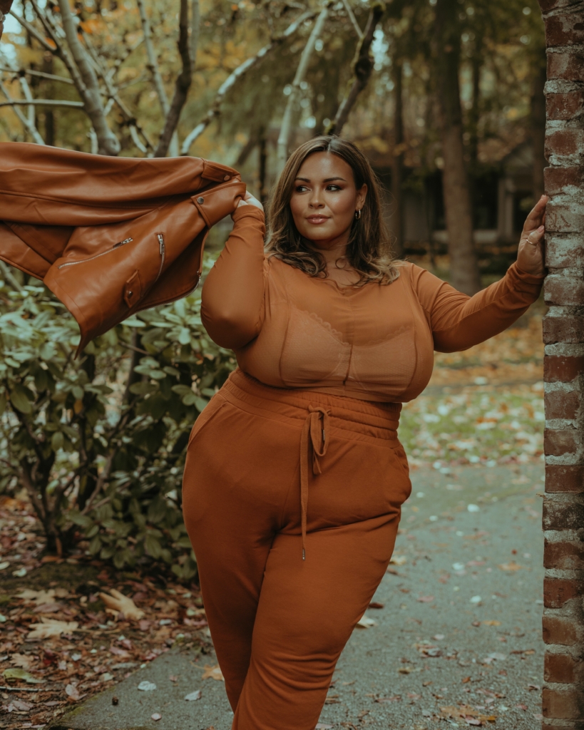 Plus size woman posing against brick wall wearing orange-tan pants and sheer top while holding matching faux leather jacket