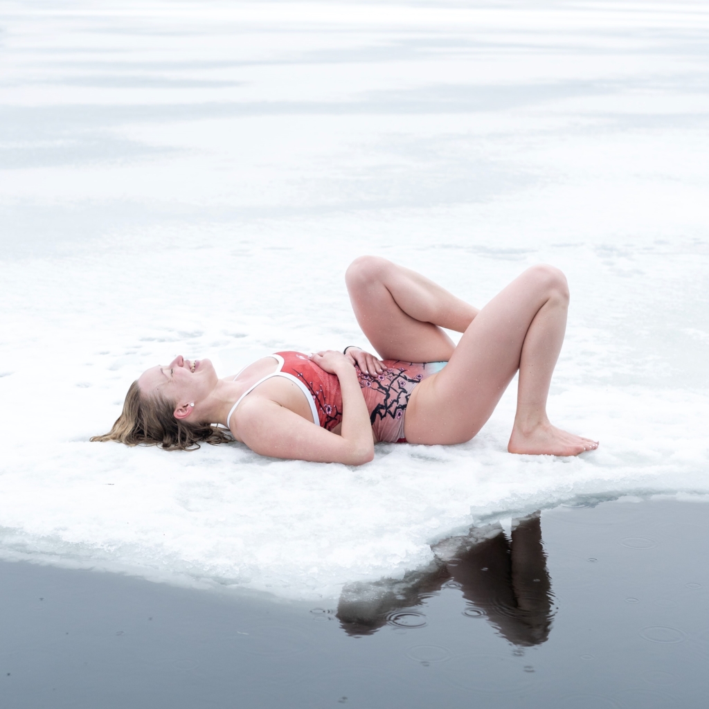Compete in your local polar bear plunge - woman lying down on snow next to freezing water and wearing a bathing suit while laughing and with wet hair