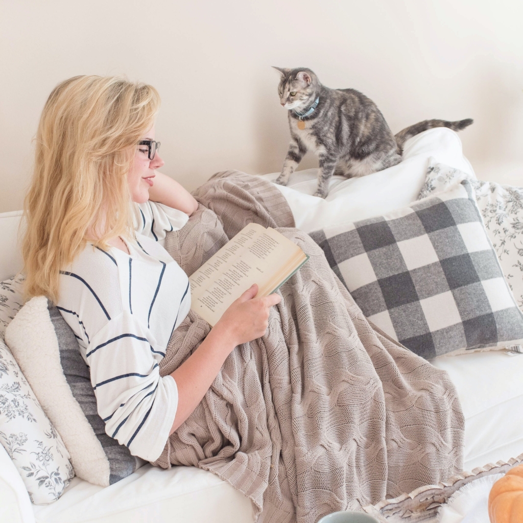 Taurus - Action and Adventure - woman sitting on couch with blankets around her and reading a book and with a cat sitting on back of couch looking at her