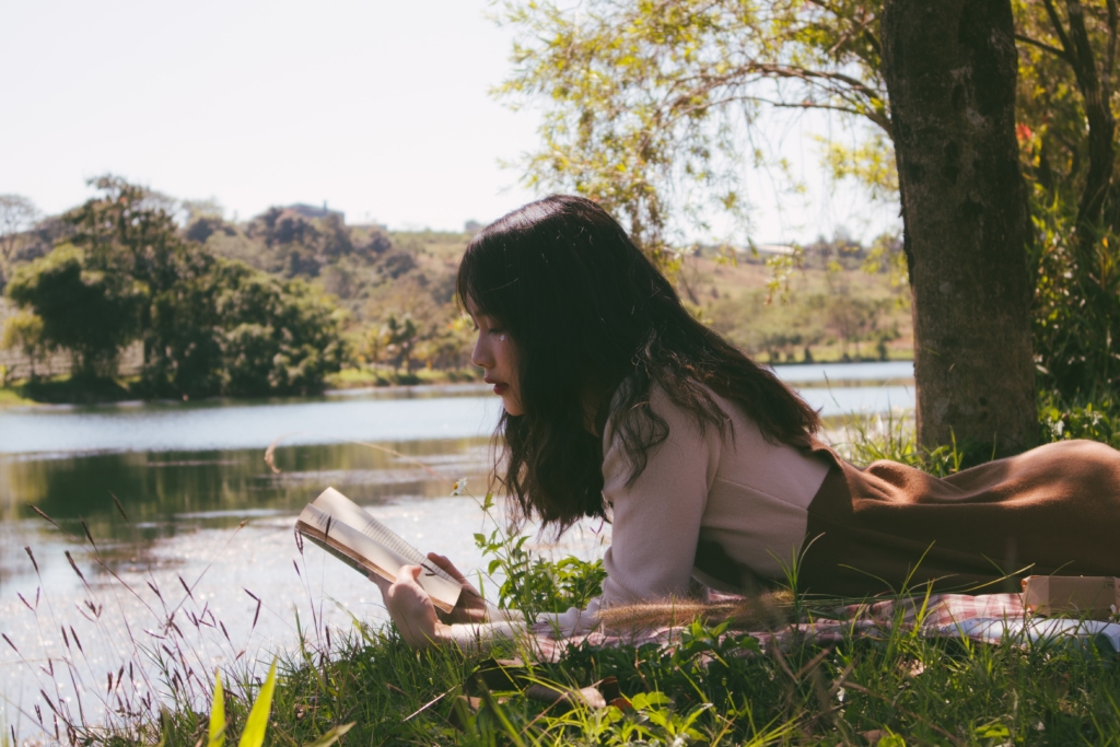 Taurus - Action and Adventure - woman lying on picnic blanket in grass next to river while reading a book
