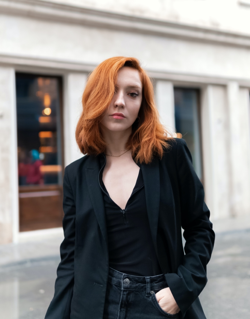 Young redhead woman standing outside wearing dark jeans, black v-neck shirt, and black blazer