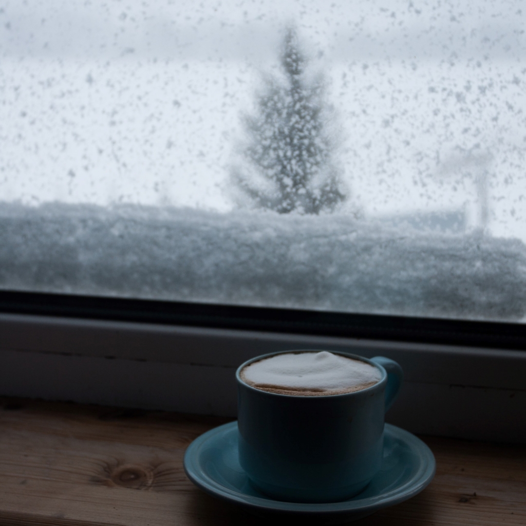 Spend time at a local coffee shop - steaming hot mug of coffee on windowsill with snowy weather outside