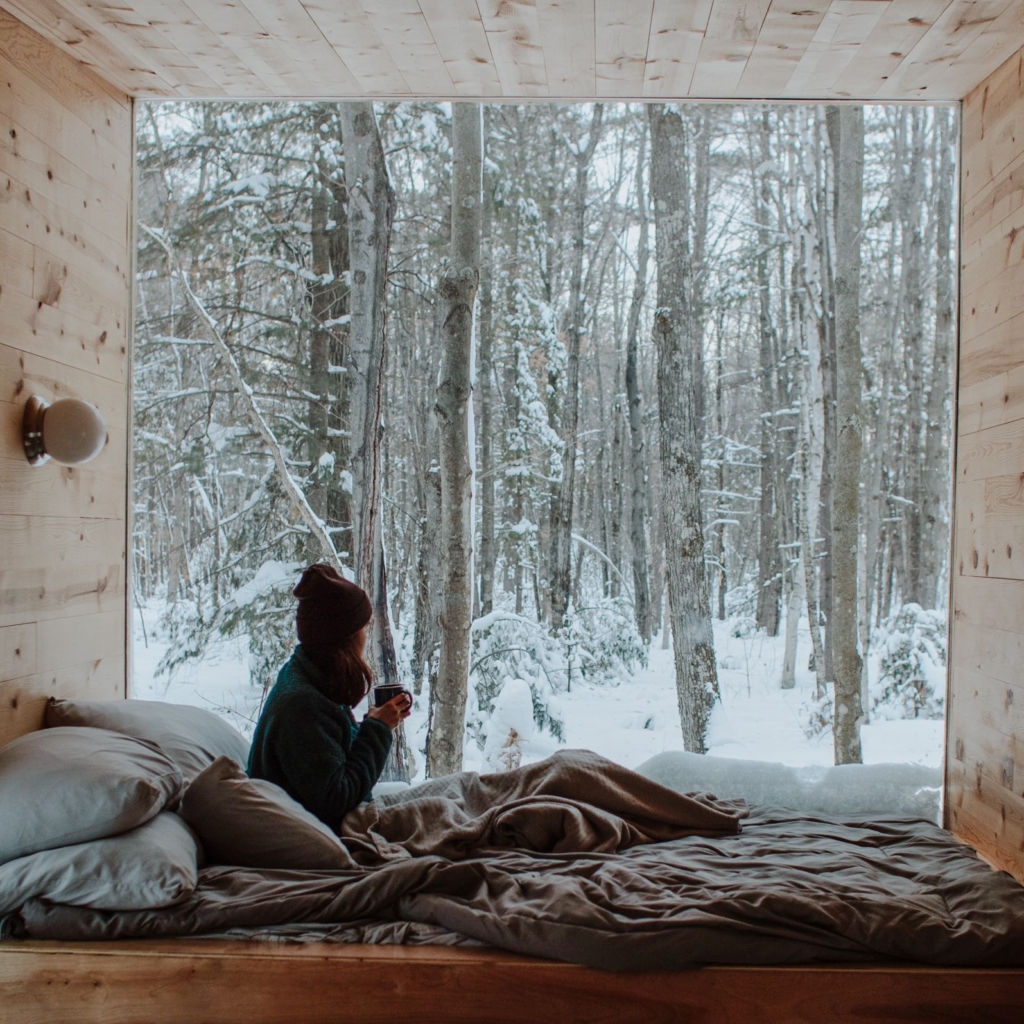 Plan a cozy cabin trip - woman snuggled in bed holding hot coffee and looking out huge window at snowy forest
