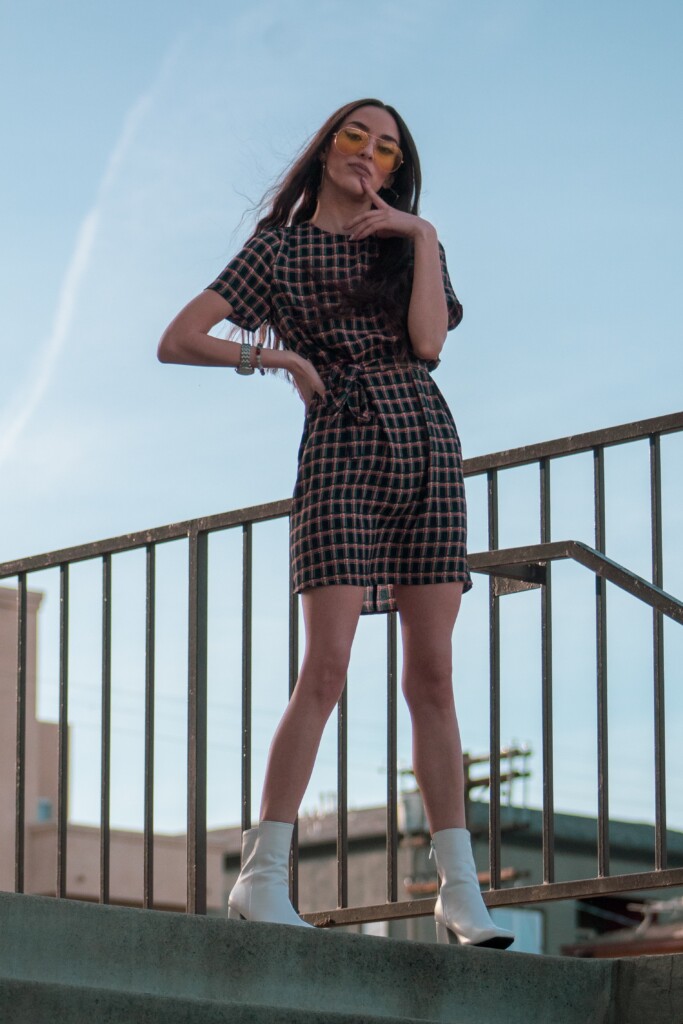 Woman standing outside posing wearing plaid dress with bow around waist and white high heel boots