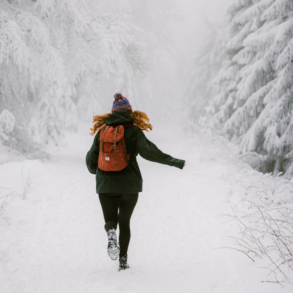 Go on a nature walk - woman running through snowy forest wearing green jacket and reddish hiking pack