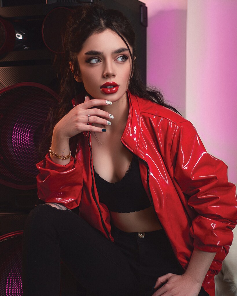 Young woman sitting wearing black bralette, black ripped jeans, and bright red faux leather jacket and bright red lipstick