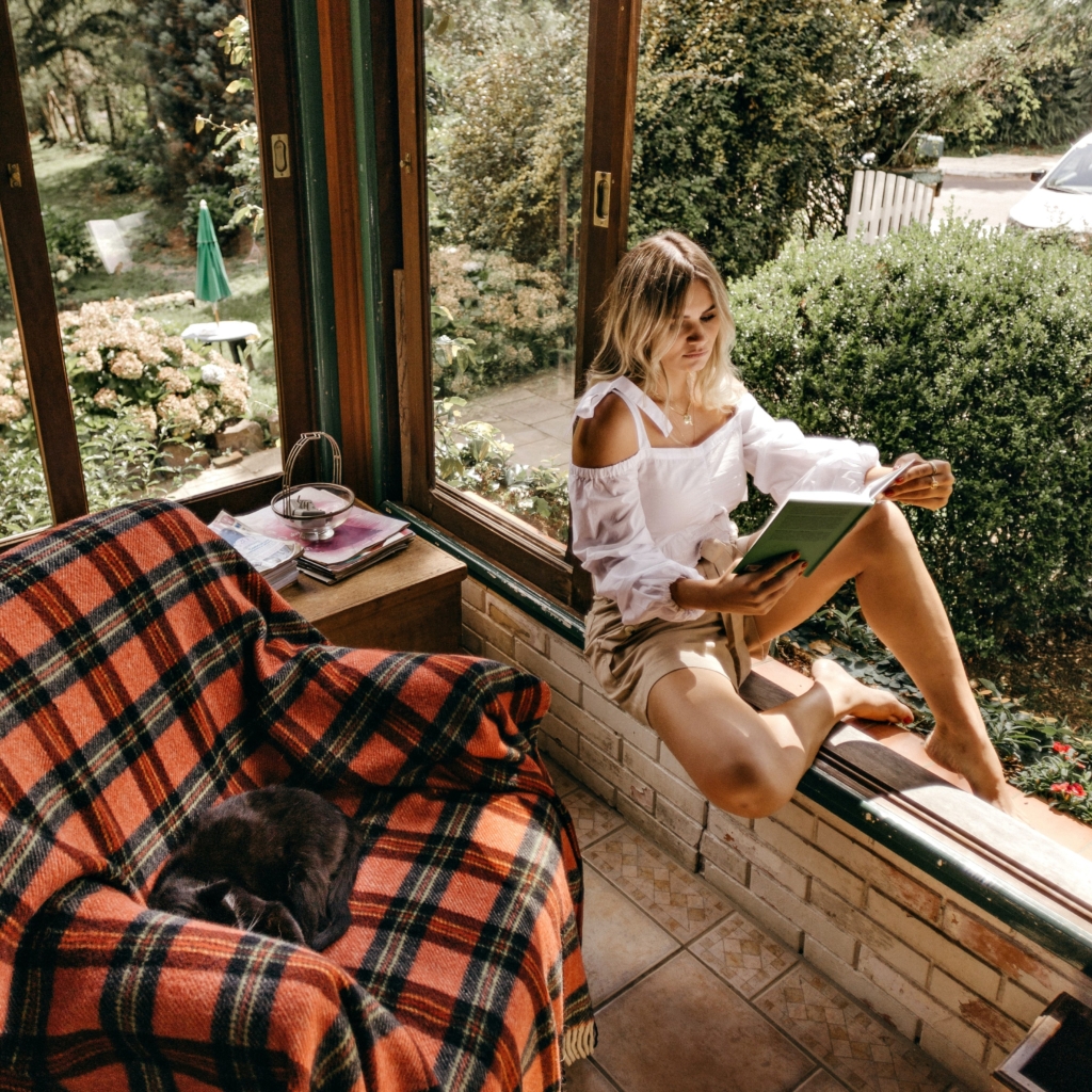 Scorpio - mystery - model sitting on open window sill reading a book in a cottage with a large beautiful garden outside and a seat inside with black cat sleeping on it